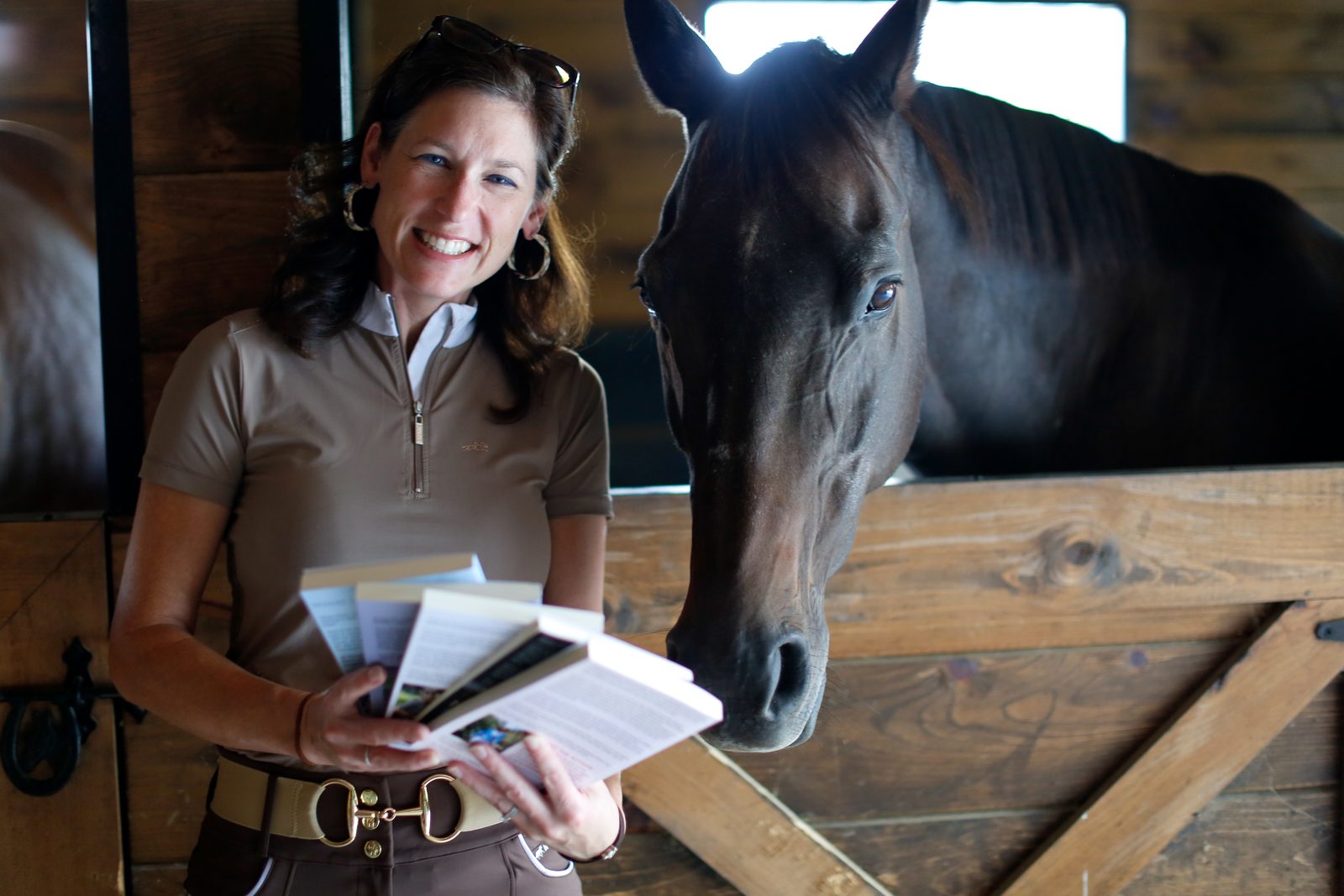 Author Laurie Berglie smiling beside her dark bay horse at a stall door, holding her books